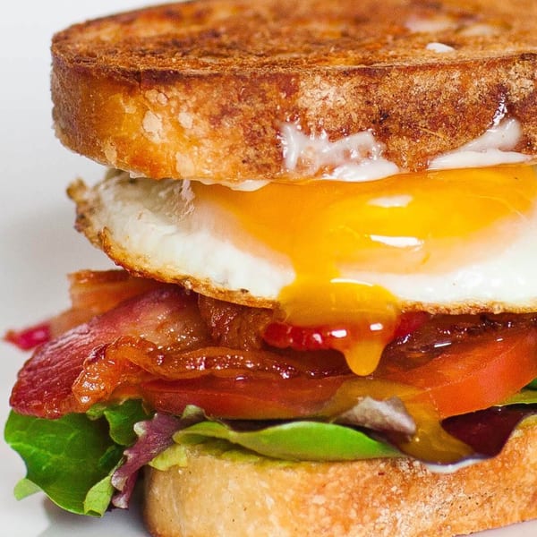 Close-up of a breakfast sandwich with a runny fried egg, bacon, tomato, and lettuce on toast.