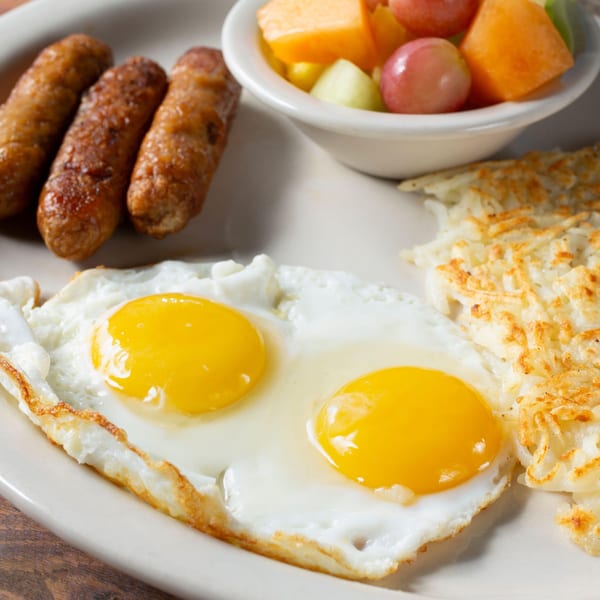 Breakfast plate with two fried eggs, three sausage links, hash browns, and a fruit cup.
