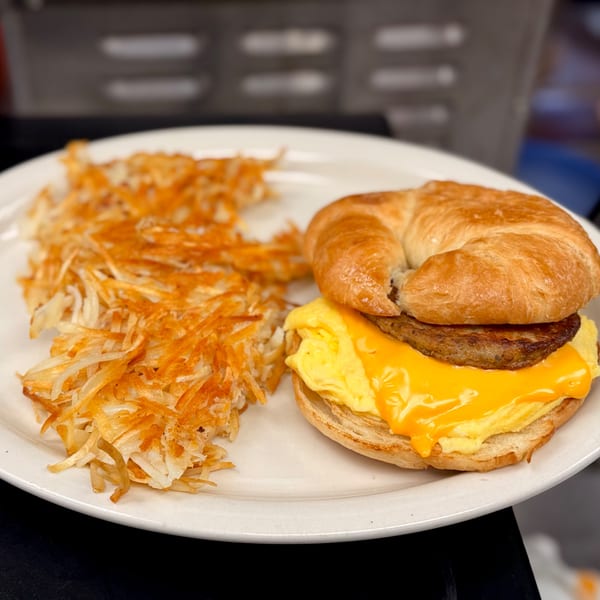 A breakfast plate with a croissant sandwich, scrambled eggs, cheese, sausage, and hash browns.