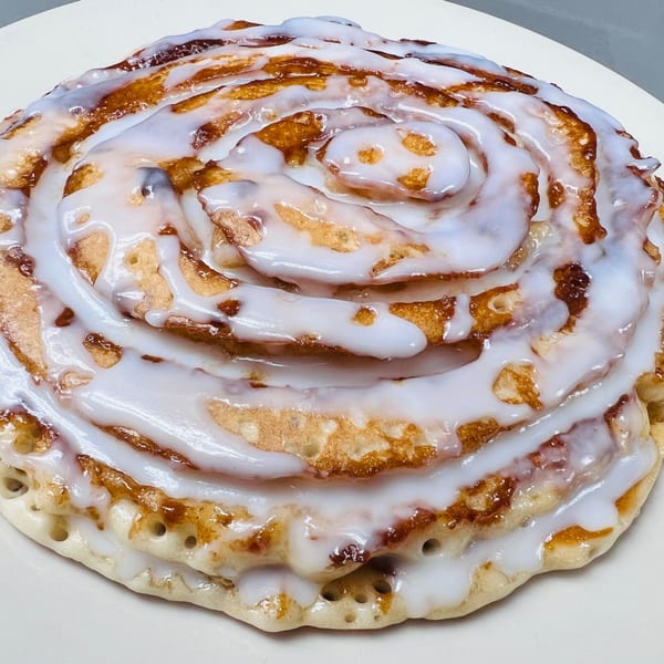 A large cinnamon roll pancake, swirled with white icing and golden-brown edges, on a white plate.