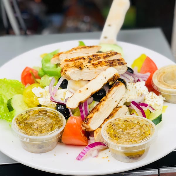 A grilled chicken salad with lettuce, tomatoes, cucumbers, olives, feta, and red onion, with dressing cups.